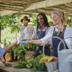 Woman shopping at a food market. Buying vegs from two women with a man in the background watching. The table has green vegs on it with a watering can.