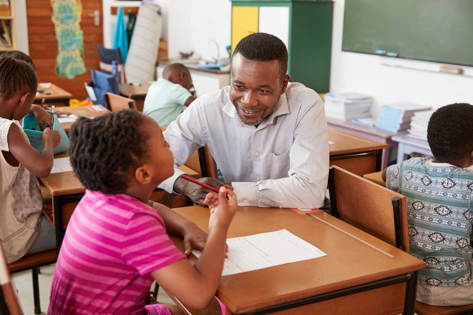 African American male teacher instructing an African American girl at a school desk with other students in the background.