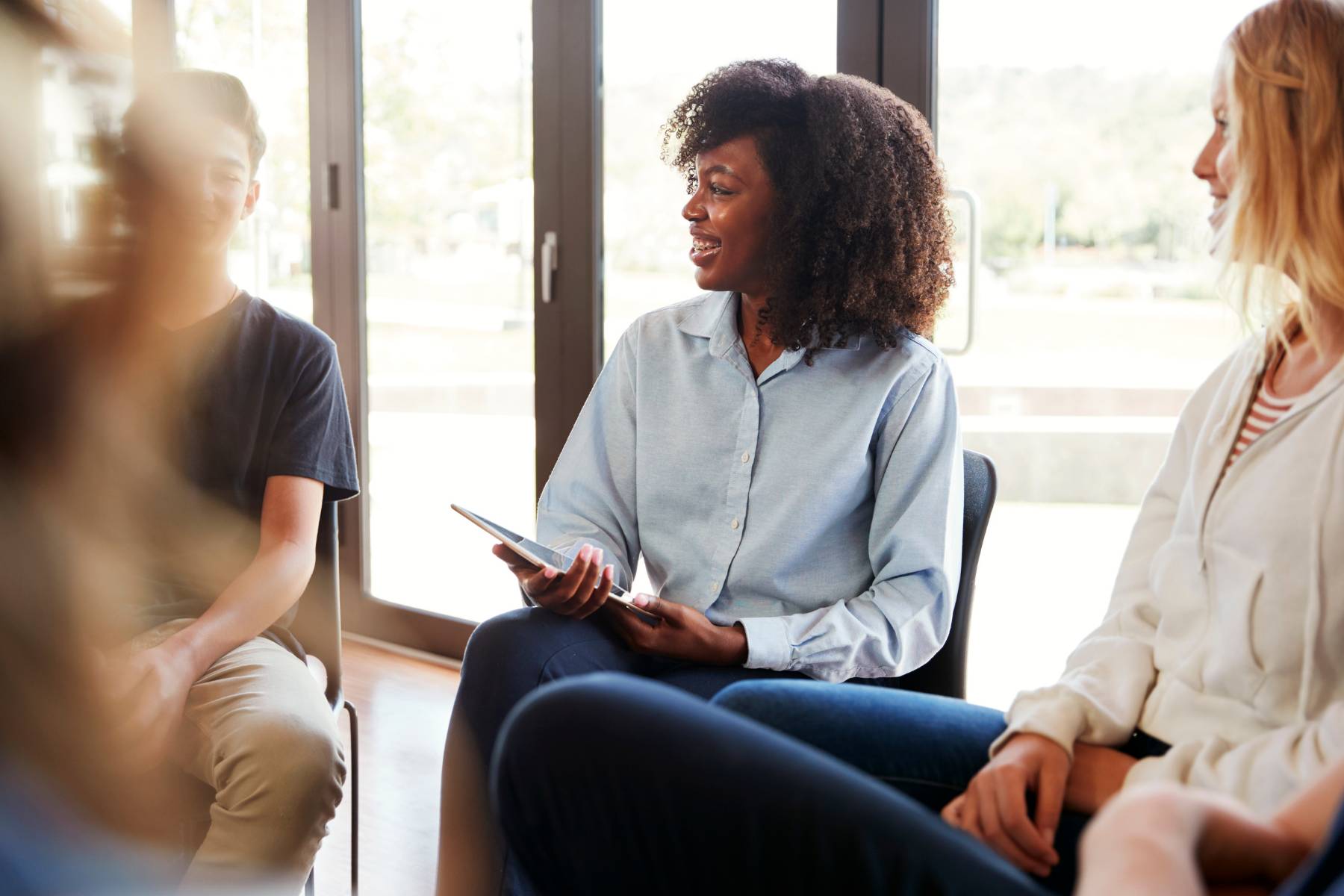 Two female and one male student sitting in front of glass doors.  One of the female student is holding an IPad