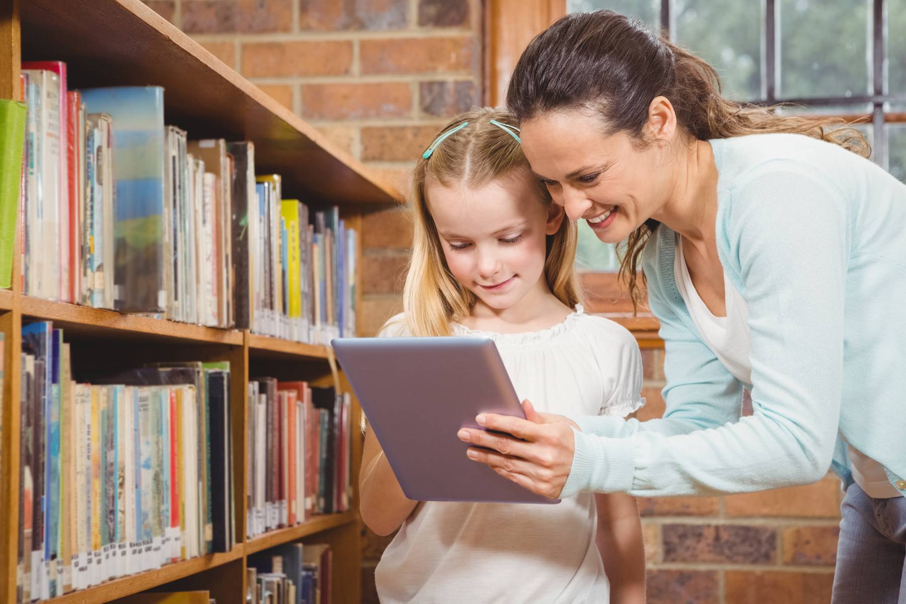 Lady showing a child information on an IPad. The two are in a library with a shelf of books to the right of them.